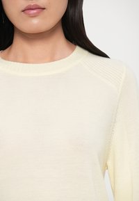 Ανοιχτό κίτρινο πλεκτό πουλόβερ με ριμπ λαιμόκοψη και υφασμάτινα ραγκλάν μανίκια με υφή. Ομαλό ύφασμα με μαλακή όψη και διακριτική λάμψη.