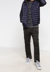 Doudoune marine à coutures horizontales, chemise rayée grise, pantalon foncé et baskets claires. La capuche est ajustable avec un cordon.