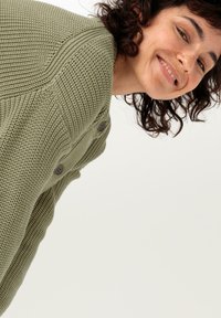 Grön stickad kardiagan med en strukturerad mönster, som har två mörka knappar och en avslappnad passform. Designen inkluderar framträdande sömmar på axlarna.