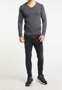 Homme portant un pull à col en V côtelé gris foncé ajusté, un jean noir texturé avec des détails aux genoux, et des baskets noires à semelles blanches.