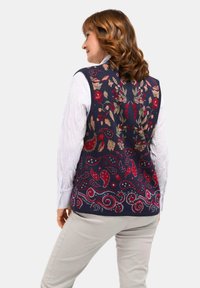 Marineblauw vest met bloemen- en paisleypatronen in rood en groen, gedragen over een wit overhemd met lange mouwen, met een relaxte pasvorm.