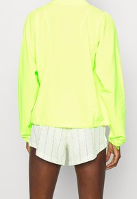 Giacca a maniche lunghe gialla neon con una texture liscia e lucida. Caratterizzata da polsini elasticizzati e orlo con coulisse, abbinata a pantaloni corti leggeri a motivi.
