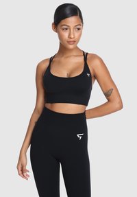 Mladá žena s tetovaním motýľa, oblečená v čiernom športovom podprsenke a legínach s vysokým pásom, sa pozerá mierne nabok na jednotnom pozadí.