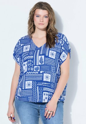 Kobieta o falowanych włosach, ubrana w niebieską i białą bluzkę w krótkim rękawie w wzory oraz niebieskie dżinsy, stojąca na jednolitym jasnym tle.