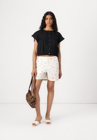 Maglia corta nera con dettagli sui bottoni e maniche a scallop, abbinata a pantaloncini bianchi con stampa floreale colorata. Borsa a tracolla marrone e sandali.