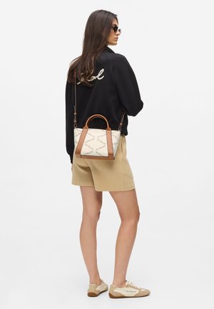 Krémová kabelka s hnedými koženými doplnkami, ozdobená textúrovaným vzorom. Nosená s čiernou mikinou a béžovými šortkami. Outfit dopĺňajú hnedé tenisky.