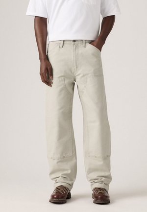 Personne portant un pantalon large beige clair, des bottes de randonnée marron, et une chemise blanche à manches courtes, avec une main dans la poche.