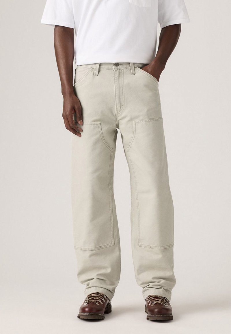 Personne portant un pantalon large beige clair, des bottes de randonnée marron, et une chemise blanche à manches courtes, avec une main dans la poche.