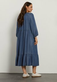 Denimmekko, jossa on kerroksellinen muotoilu, pitkät puff-hihat ja pyöreä pääntie. Yhdistetty valkoisiin tennareihin harmaalla lattialla.