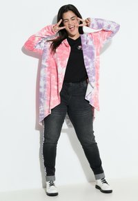 Larga chaqueta de punto con estampado de tie-dye en rosa y morado, sobre una camiseta negra, combinada con jeans oscuros y botas blancas con cordones.