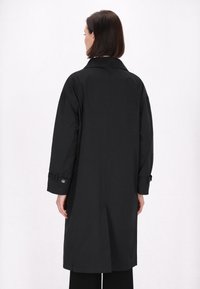 Langer schwarzer Trenchcoat mit geradem Schnitt, großem Kragen, hochgekrempelten Ärmeln und Knopfdetails an den Ärmeln. Glatter Stoff und minimalistisches Design.