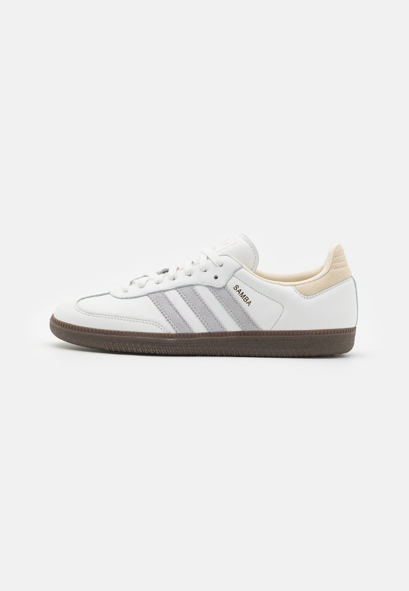 White leather sneaker with three gray stripes, round toe, lace-up front, and brown rubber sole. Back heel features a beige accent.