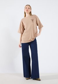 Camiseta beige de gran tamaño con mangas cortas y un estampado gráfico. Combinada con jeans de pierna ancha azul oscuro y botas verdes oliva de punta.