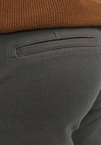 Dunkelgraue Textilstoff-Hosen mit einer flachen Gesäßtasche, kombiniert mit einem braunen Strickpullover, der ein sauberes, einfaches Design zeigt.