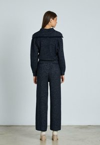 Kvinna som vänder bort ansiktet, iklädd en mörkblå ribbad stickad tröja och vida byxor, tillsammans med beiga klackstövlar mot en enfärgad bakgrund.