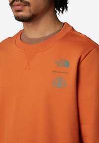 Orange bomullströja med rund halsringning, ribbade ärmar och tryckta logotyper i turkos på bröstet och axeln.