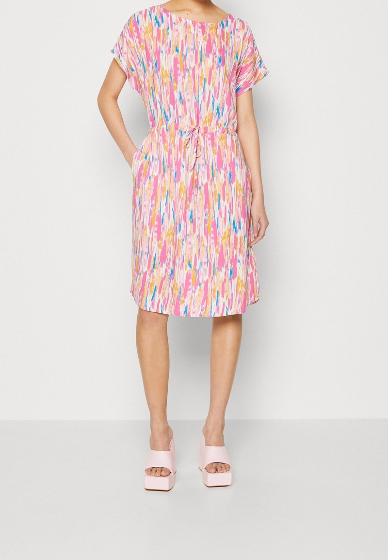Femme portant une robe mi-longue à manches courtes avec un motif abstrait rose, bleu et orange, associée à des sandales compensées rose clair.