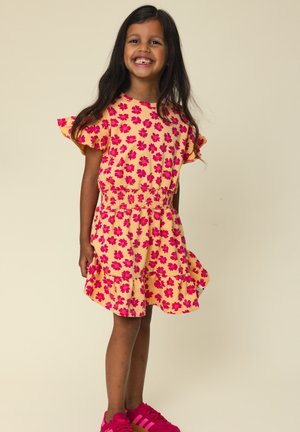 Glimlachend meisje met lang donker haar draagt een gele jurk met een roze bloemenpatroon en felroze sneakers, staand tegen een lichte achtergrond.