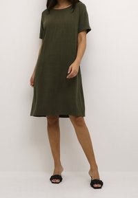 Vestido verde oliva de mangas cortas con cuello redondo, de forma A, hecho de un tejido texturizado, que termina por encima de la rodilla. Llevado con sandalias negras.