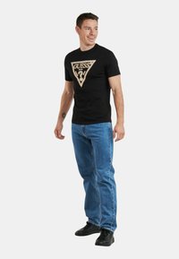 Camiseta de algodón negra con un gráfico triangular beige que presenta el logo "GUESS"; combinada con jeans azules y zapatillas negras.