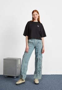 Black cropped t-shirt with a logo, light blue cargo jeans with multiple pockets, paired with beige sneakers. Gray rolling storage box nearby.