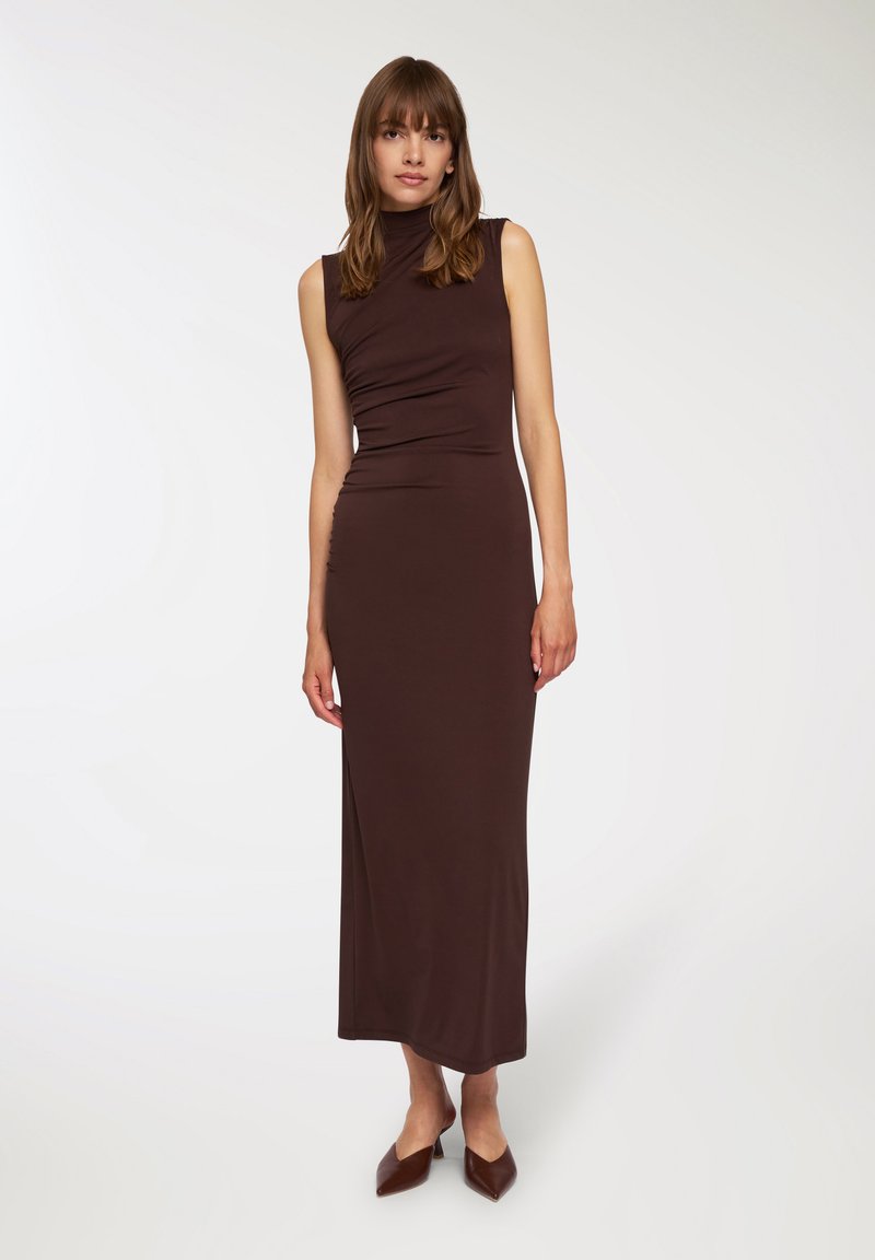 Dopasowana, bezrękawnikowa brązowa sukienka z wysokim dekoltem i marszczeniami, wykonana z gładkiej tkaniny. Nosi się ją z brązowymi, spiczastymi obcasami.