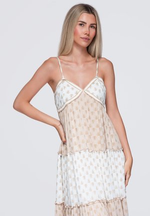 Lyshåret kvinde iført en ærmeløs lagdelt kjole med tynde stropper og beige blomsterprint, poserer med den ene hånd på hoften mod en neutral baggrund.
