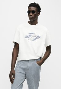 Άνδρας με στρογγυλά μαύρα γυαλιά ηλίου, λευκό t-shirt με το λογότυπο Dickies, και μπλε και λευκή ριγέ παντελόνια, με το χέρι στην τσέπη, σε απλό ανοιχτόχρωμο φόντο.