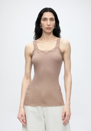 Mujer con largo cabello negro lleva una camiseta sin mangas ajustada de canalé con ribete de encaje en beige y pantalones blancos holgados sobre un fondo liso.