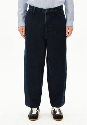 Person trägt lockere dunkelblaue Jeans, hellblaues Hemd und schwarze Sneakers mit weißen Schnürsenkeln und steht vor weißem Hintergrund.