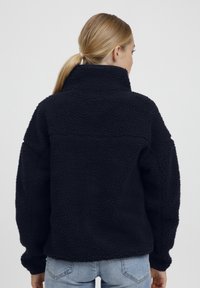 Svart fleece-genser med høy krage, teksturert overflate, senkede skuldre og løs passform. Bæres med lyseblå jeans.