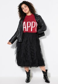 Roter Strickpullover mit dem Aufdruck "HAPPY", schwarze Lederjacke und gestufter schwarzer Tüll-Midirock, kombiniert mit schwarzen Nieten-Stiefeletten.