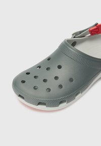 Graue Gummischuhe mit Belüftungsöffnungen, strukturiertem Riemen und weißer Sohle. Sie verfügen über ein flaches Design und eine abgerundete Zehenpartie für optimalen Komfort.