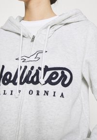 Šedá mikina na zip s nášivkou "Hollister" v námořnické modři, bílou podšívkou a nastavitelnou kapucí se šňůrkou.