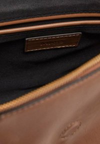 Brown leather bag interior with black fabric lining, featuring a zipped pocket, brown stitching, and a small brand label.
