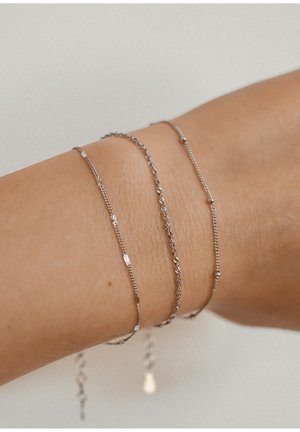 Three delicate silver bracelets on a wrist: one with small flowers, one twisted, and one with tiny beads. Sleek, fine designs.
