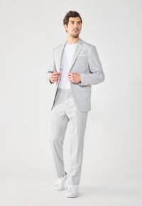 Hellgrauer Anzug mit taillierter Passform, einreihiger Blazer, passende Hose und weiße Sneakers. Mit einem Detail in Form eines Einstecktuchs.