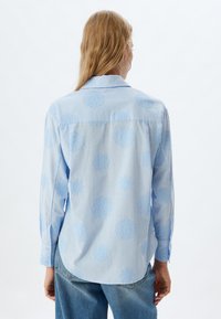 Camisa de manga larga azul clara con patrones florales bordados, cuello de tipo botón y dobladillo redondeado. La tela es texturizada y suave.