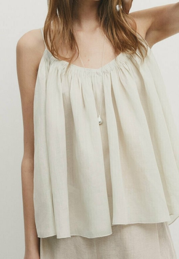 Femme portant un haut ample sans manches beige clair avec des bretelles fines et un collier pendentif sur un fond neutre.