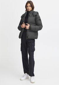Puffjakke i mørk grå, med høy krage, glidelås og quiltet design. Kommer med marineblå cargo-bukser og hvite sneakers.