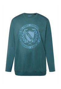 Maglietta a maniche lunghe di colore teal, realizzata in tessuto morbido, con una grafica circolare che presenta la scritta "Champion Spirit" e un'icona a forma di scudo, datata "1997".