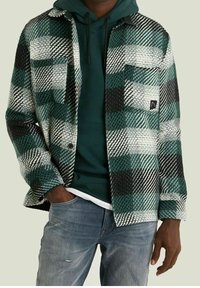 Man i blekta blå jeans, mörkgrön huvtröja och en grön, svart och vit rutig skjorta med knappar och fickor.