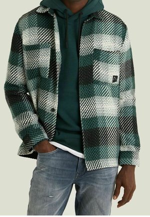 Homme portant un jean bleu décoloré, un sweat à capuche vert foncé et une veste à carreaux vert, noir et blanc avec des poches.