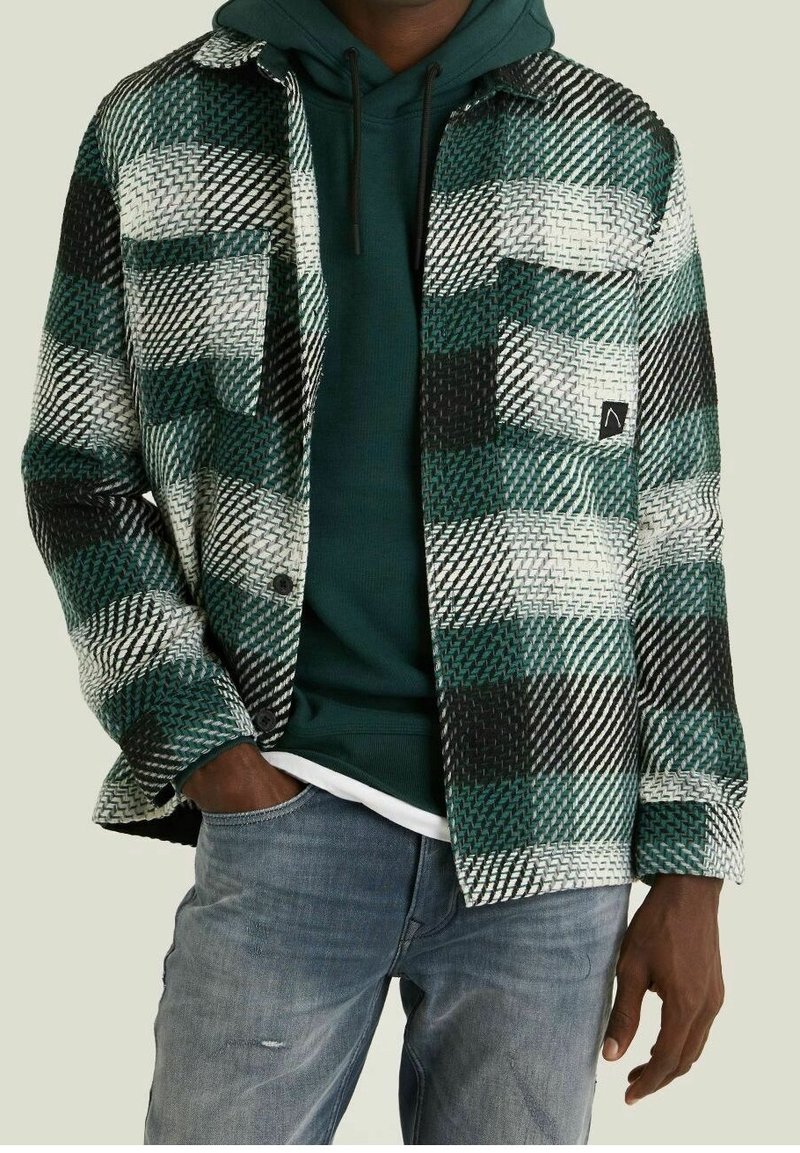 Man i blekta blå jeans, mörkgrön huvtröja och en grön, svart och vit rutig skjorta med knappar och fickor.