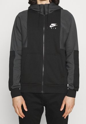 Man in een zwart en donkergrijs Nike Air hoodie met rits, voorzakken en trekkoordkap, staand met ontspannen handen.