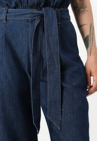 Donkerblauwe spijkerbroek met een hoge taille, voorzien van een gestrikt riem, zichtbare stiksels in contrasterend oranje en een ontspannen pasvorm.