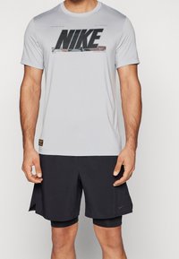T-shirt de sport gris avec un logo "NIKE" noir sur la poitrine, associé à des shorts de sport noirs. Le tissu semble lisse et léger.