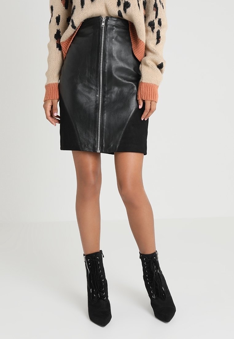 Femme portant une mini-jupe en cuir et suède noir avec une fermeture éclair à l'avant, associée à un pull imprimé léopard et des bottines noires à lacets.