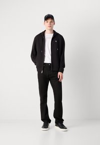 Sudadera negra con cremallera y cordones, camiseta blanca con logo, jeans negros y zapatillas negras con suelas blancas, de pie contra un fondo sencillo.