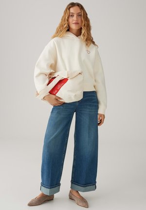 Junge Frau steht und trägt einen cremefarbenen Hoodie mit kleinem Herz, blaue weite Jeans, beigen Ballerinas und hält eine beige Einkaufstasche mit rotem Streifen und der Aufschrift "Love Edition".